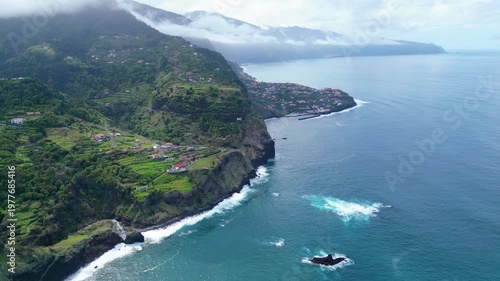 Aerial view of Madeira's dramatic coastline, featuring lush terraced hillsides, Boaventura villages, and the vibrant Atlantic Ocean under a cloudy sky.