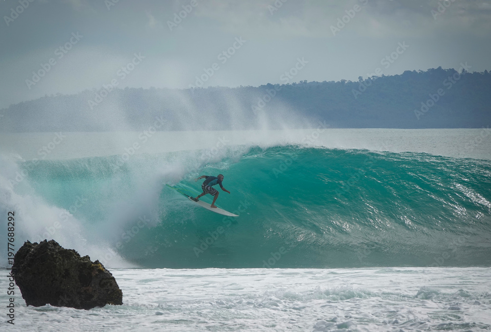 Naklejka premium Male surfer rides a fast and hollow turquoise wave with offshore mist. Exposed sharp rock in the foreground shows danger of surfing reef breaks in tropical paradise of Mentawai Islands in Indonesia.