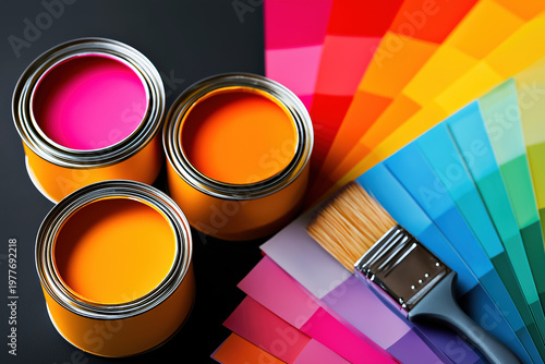 Top view of three open paint cans in orange red, purple with a paintbrush and rainbow color swatch fan on dark background