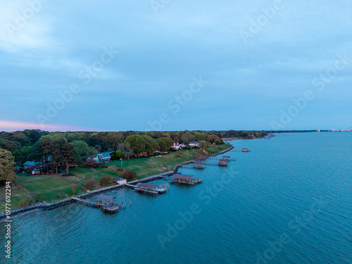 Foto Aerial view of Newport News coastline along the James River, featuring a row of private wooden piers and boat houses along the grassy shoreline at dusk