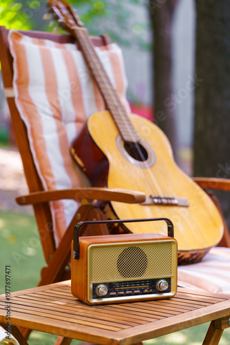 Cozy outdoor relaxation space with wooden garden chair, acoustic guitar and vintage radio placed on small table. Peaceful backyard leisure scene for music, rest and summer recreation in green garden.
