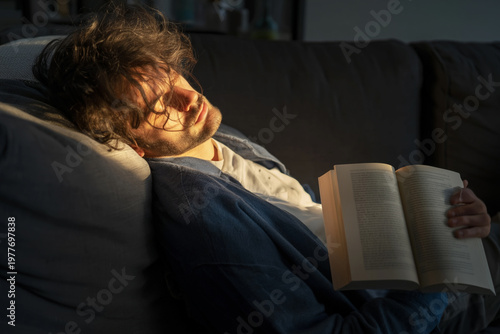 Young man peacefully napping on a cozy sofa with a book in hand during the day