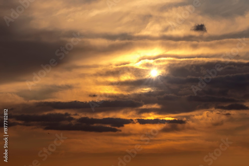 Zachmurzone, baśniowe niebo o zachodzie słońca z warstwami ciemnych i jasnych chmur tworzy dramatyczny efekt. Słońce jest częściowo przesłonięte przez chmury, rzucając ciepłe, złote światło na scenę. 