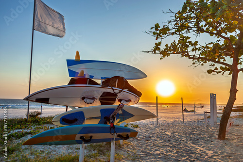 stack of surfboards on a rack at Praia do Pepê, Rio de Janeiro, during a golden sunset. The beach scene features white sand, a flag, and a tree, capturing the vibrant kitesurfing culture