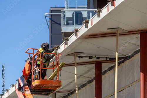 Construction Worker in High Visibility Gear Operating Orange Aerial Boom Lift to Work on Building Canopy
