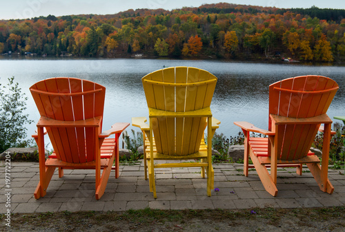 Two orange and one yellow Muskoka chairs are arranged on a stone paver patio overlooking a calm Ontario lake and a rolling hillside of peak autumn foliage on an overcast fall day. The mix of orange