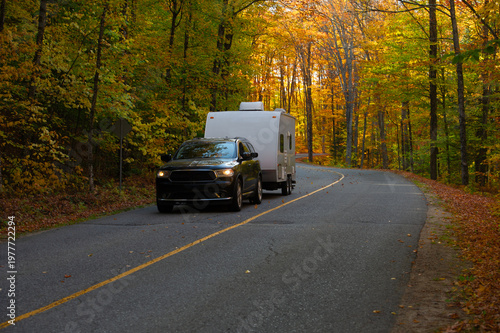 Cuadro en lienzo A black SUV towing a white travel trailer rounds a winding forested road through peak autumn foliage in Ontario, Canada, with headlights on