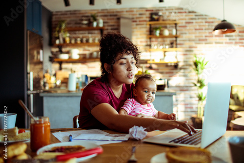 Mother working on laptop with baby at home kitchen table