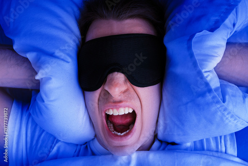 Foto Angry man in a sleep mask screams while covering his ears with pillows in bed