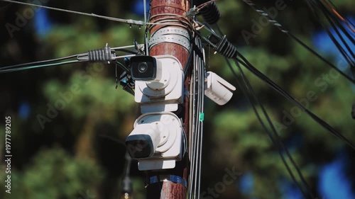 Close up of a utility pole with surveillance cameras, wires, and hanging lights against a blue sky