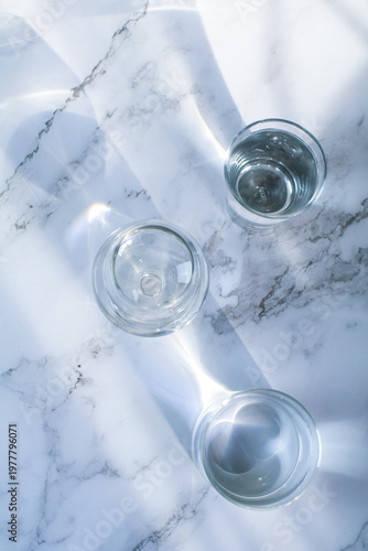 Glass of water under sunlight creating abstract shiny reflection on marble floor. Minimal aesthetic top view summer lifestyle.