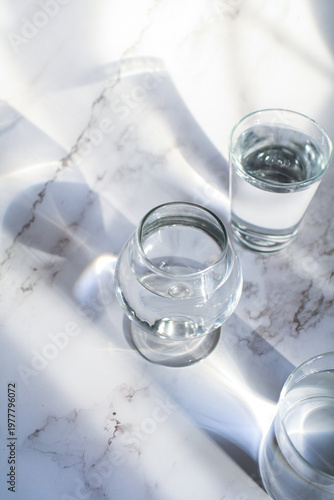 Glass of water under sunlight creating abstract shiny reflection on marble floor. Minimal aesthetic top view summer lifestyle.