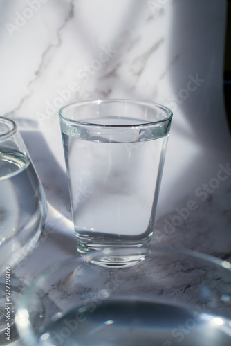 Glass of water under sunlight creating abstract shiny reflection on marble floor. Minimal aesthetic top view summer lifestyle.