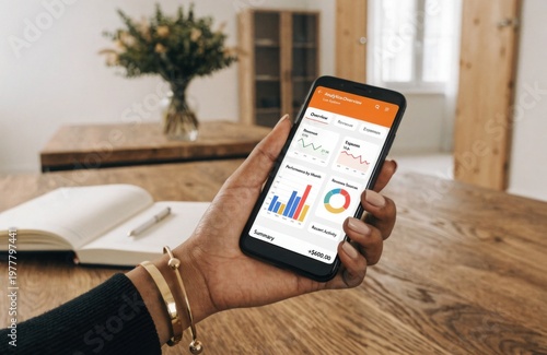 Professional business woman�s hand checking data charts on smartphone while working remotely at a bright minimalist wooden desk with natural light. (139 characters)