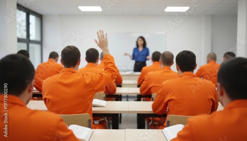 Male inmates in orange jumpsuits attend a classroom lecture, with one raising his hand to ask a question to the female teacher.