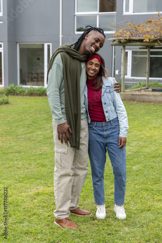 Indian and African American couple standing on grassy lawn by gray building, wearing denim jacket