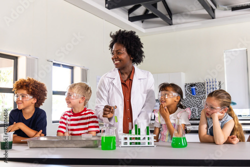 Adult female educator in lab coat leading chemistry demo at lab bench, four children in goggles