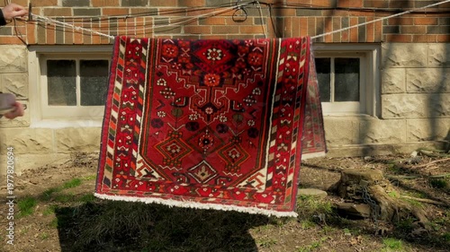 Persian rug being beaten with broom to remove dust during spring cleaning, hanging on clothesline against brick building exterior, Toronto, Canada.