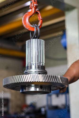 Close up of a heavy duty steel helical gear and splined shaft assembly suspended by an industrial crane hook in a workshop manufacturing facility.