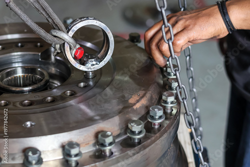 Close-up photo of a mechanic's hand inserting bolts into a heavy metal industrial gear housing for transmission assembly in a workshop, focusing on manual installation by fingers.
