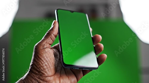 A dark-skinned hand holding a smartphone with a green screen against a blurred green background with white outline, technology concept in a realistic photographic style