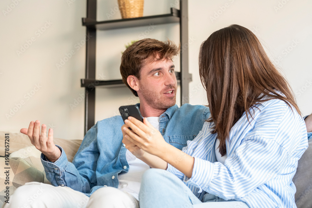Obraz premium Young couple sit on a living room sofa, engaged in a tense discussion over a smartphone, showing frustration and a communication breakdown during an emotional domestic moment.