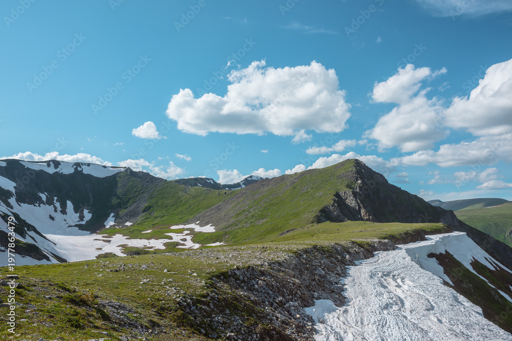 Naklejka premium Scenic sunlit view along grassy ridge with white snow cornice to green hill top with rocky sheer crags in bright sun under lush clouds in blue sky. Vivid alpine scenery in high mountains in sunny day.