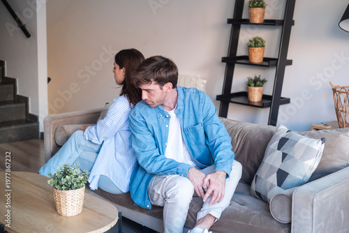 Foto Tense young couple sit back to back on a sofa in a modern living room after an argument, showing distance, frustration and emotional disconnect in a domestic setting