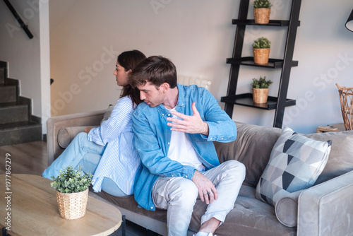 Photos A young couple sits back-to-back on a sofa after an argument, showing tension and emotional distance in a contemporary living room setting with neutral decor and casual clothing