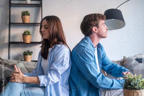 Obraz na plátně Young man and woman sit back-to-back on a sofa in a living room, showing tension and emotional distance after a disagreement