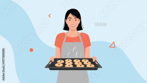 Smiling young woman wearing apron holding tray of freshly baked homemade cookies in warm kitchen environment.