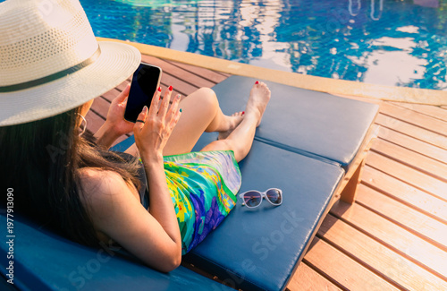 Schilderij op canvas Asian woman with hat and swimsuit sit on sunbed at poolside and using smartphone on summer vacation by swimming pool