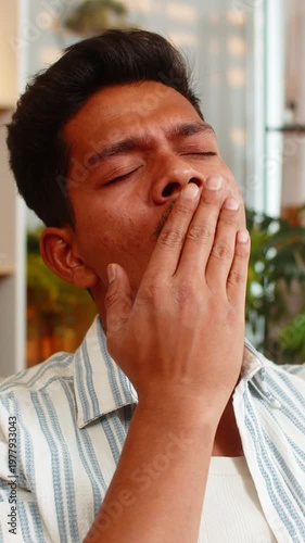 Indian man on home sofa rubs tired eyes and yawns with hand near face during late sleepy evening rest. Arabian boy on home sofa feels drowsy and worn out after long work and quiet home rest indoors.