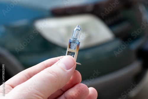 A person holds a new car headlight bulb close to vehicle headlights. The setting is outdoors in daylight. The vehicle is parked nearby.