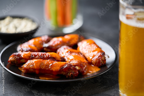 bbq buffalo chicken wings in a plate, with beer, carrot and celery sticks on a dark table.