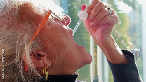 Side profile close-up of a senior woman using a glass dropper to take oral liquid medication. Concept of elderly healthcare, natural supplements, and daily wellness routine at home
