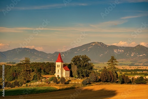Idyllic countryside landscape in the Turiec Basin near Martin and Turčianske Teplice, Slovakia. Small rural church with red roof surrounded by fields and trees, with Velka Fatra mountains