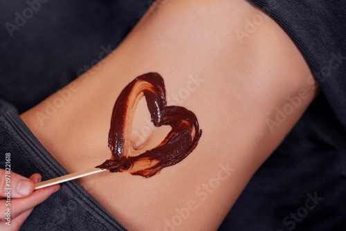 Female figure with smooth skin, applying chocolate body mask in heart shape on abdomen, showcasing beauty treatment and self-care ritual for relaxation and wellness