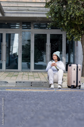 Woman in hoodie sitting curb by glass entrance holding to-go cup with wheeled suitcase, copy space