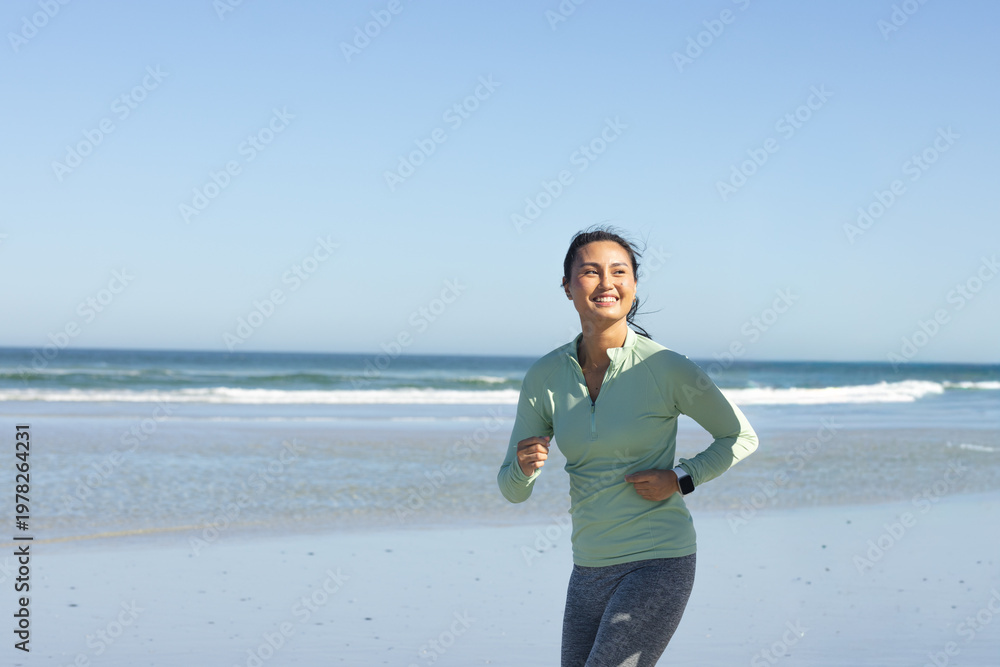 Naklejka premium Mid-adult Asian woman jogging along wet sand by sea in green top checking smartwatch, copy space