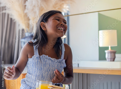 Female child laughing, gesturing at dining table in light blue dress with orange juice and pancakes