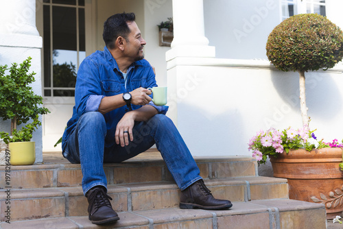 Middle-aged Asian man sitting on porch steps in denim shirt holding light green mug, looking left