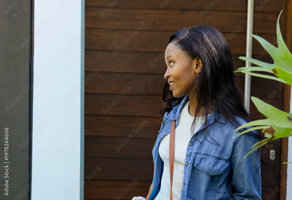 Naklejka premium African American woman standing by wood-paneled door, wearing denim, holding bottle, copy space