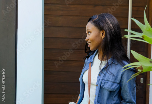African American woman standing by wood-paneled door, wearing denim, holding bottle, copy space