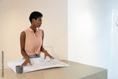 African American woman reviewing blueprints on counter in studio, wearing blouse with gray mug