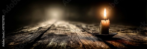A single candle burns brightly on an old wooden surface, casting warm light across the textured grain. The dark background emphasizes the flame's glow and creates deep shadows around it.