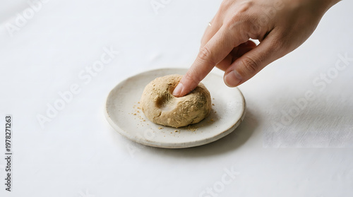 A hand gently pressing a finger into a soft, powdery round snack on a rustic ceramic plate, leaving a delicate indent.