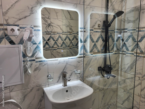 The bathroom has a lighted mirror, a hairdryer, a sink, and a glass partition with a shower.