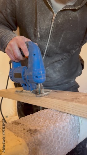 Close-up of a craftsman’s hand using a jigsaw to cut and shape wood. Detailed woodworking process in a workshop setting.