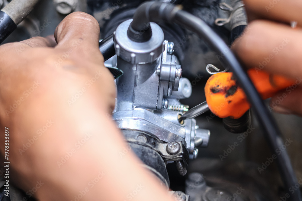 Obraz premium ​A mechanic uses an orange-handled screwdriver to precisely adjust the fuel-air mixture screw on a motorcycle carburetor in a workshop, ensuring optimal engine performance and smooth idling.